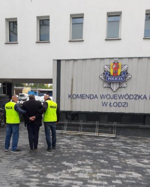 dwaj policjanci konwojują podejrzanego o łapownictwo, zboku logo łódzkiej Policji z napisem Policja, Komenda Wojewódzka w Łodzi