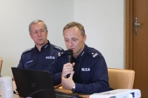 uczestnicy narady dwaj policjanci siedzą przy stole jeden trzyma mikrofon, do którego mówi