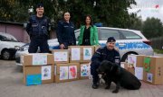 Na zdjęciu widoczni policjanci, pies służbowy oraz przedstawicielka schroniska z Tychów. Wszyscy stoją przy rozłożonych kartonach z darami dla zwierząt, w tle widoczny radiowóz.&quot;&gt;