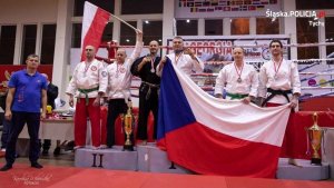 Na zdjęciu widoczne podium, na którym stoją mężczyźni po dwóch na każde z miejsc na podium. Po lewej stronie zdjęcia, na drugim miejscu widoczni Polacy, jeden z nich trzyma uniesioną w górę flagę Polski