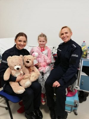 policjantki siedzą przy łóżku dziewczynki w szpitalu, która trzyma przekazane maskotki