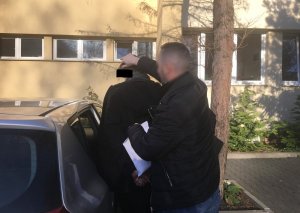 Na zdjęciu parking policyjny, policjant pilnuje zatrzymanego, pomaga mężczyźnie do wsiąść do radiowozu nieoznakowanego