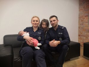 policjantka trzyma na rękach niemowlę obok siedzi policjant a zza ich pleców wygląda dziewczynka