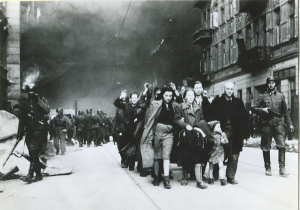 czarno-białe zdjęcie przedstawiające grupę Żydów idących w grupie przez miasto oraz żołnierzy niemieckich z karabinami