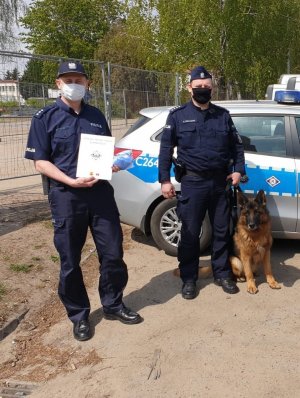dwaj policjanci stoją obok radiowozu policyjnego, jeden z nich trzyma na smyczy psa o imieniu Harry, drugi policjant trzyma podziękowanie
