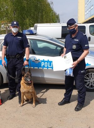 dwaj policjanci stoją obok radiowozu policyjnego, jeden z nich trzyma na smyczy psa o imieniu Taj, drugi policjant trzyma podziękowanie