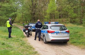 dwaj policjanci, auto, którym uciekał kierujący i policyjny motocykl na leśnej drodze