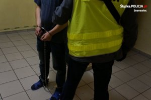 zatrzymany mężczyzna z założonymi kajdankami na rękach i nogach obok niego stoi policjant