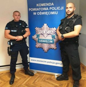 dwaj policjanci z Oświęcimia stoją na tle banera z napisem komenda Powiatowa Policji w Oświęcimiu, pod napisem stylizowana gwiazda policyjna