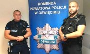 dwaj policjanci z Oświęcimia stoją na tle banera z napisem komenda Powiatowa Policji w Oświęcimiu, pod napisem stylizowana gwiazda policyjna