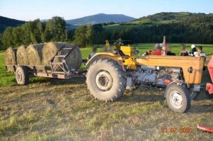 traktor rolniczy z podczepioną przyczepą, na której znajdują się cztery bale słomy