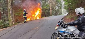Na zdjęciu na pierwszym planie policjant na motorze w tle widoczna droga i strażak gaszący w rowie palący się pojazd marki Audi