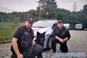 dwaj policjanci z psem obok radiowozu