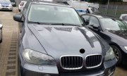 odzyskany przez policjantów samochód marki BMW X5, widok przodu pojazdu, który jest w kolorze ciemnym, w tle policyjne radiowozy i budynki