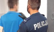 Policjant w mundurze trzyma za ramię drugiego mężczyznę, który ma założone kajdanki na ręce trzymane z tyłu.&quot;&gt;