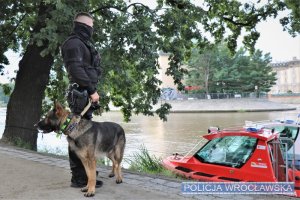 Na zdjęciu funkcjonariusz z psem służbowym, stojący koło łodzi ratunkowej nad Odrą.