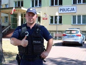 St. sierż. Maciej Kąkol w mundurze z kciukiem uniesionym do góry stoi przed budynkiem Komendy Powiatowej Policji w Otwocku. W tel widać policyjny radiowóz