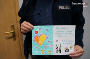 Laurka wykonana przez dzieci, na której znajduje się następująca treść podziękowania: &quot;Dziękujemy Dzielnicowym za codzienne odwiedziny troskę oraz życzliwy uśmiech&quot;. Laurkę trzyma policjant, którego widać jedynie fragment