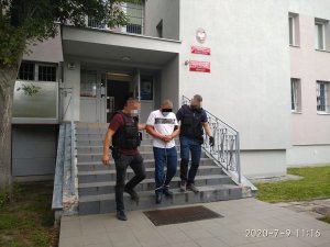 Dwóch policjantów (po bokach) wyprowadza z komisariatu w Bydgoszczy jednego z podejrzanych w kajdankach na rękach i na nogach (w środku), widać ich przodem, twarze są zamazane