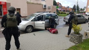Samochód z otwartymi drzwiami, podejrzany leży na brzuchu w kajdankach na ziemi, obok stoi czterech funkcjonariuszy policji