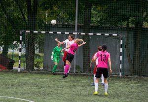 Boisko sportowe, na tle bramki dwie piłkarki walczą o piłkę w powietrzu, dwie stojące tyłem obserwują przebieg akcji, w bramce stoi bramkarka.