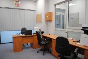 Wnętrze Posterunku Policji w Grodzisku - w pomieszczeniu znajdują się trzy biurka i trzy krzesła, na jednym z biurek leży laptop, na ścianie wisi godło Polski
