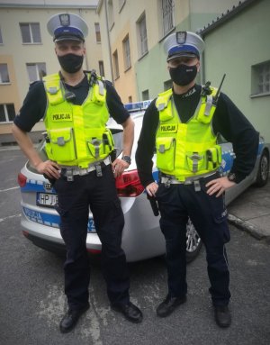 dwaj policjanci ruchu drogowego ubrani w żółte odblaskowe kamizelki z napisem: Policja , stoją przed policyjnym radiowozem