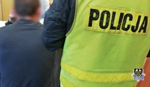 Zdjęcie przedstawia zatrzymaną osobę i stojącego obok tyłem policjanta w odblaskowej kamizelce z napisem policja