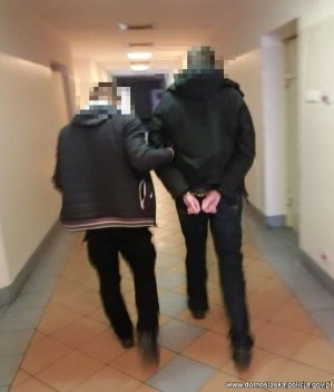 Na zdjęciu policjant w cywilnym ubraniu prowadzi korytarzem komendy Policji skutego w kajdanki zatrzymanego