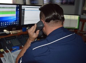 policjant dyżurny siedzi przy biurku i rozmawia przez telefon, przed nim na biurku stoją 3 ekrany komputerów