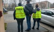 Zdjęcie jest zrobione na dziedzińcu Komendy Miejskiej Policji w Łodzi, widzimy na nim zatrzymanego ubranego w ciemną kurtkę, dżinsy. Jest wysoki, szczupłej budowy ciała, ma ciemne włosy. Ręce ma skute kajdankami. Po jego lewej stronie trzyma go policjant ubrany w żółtą kamizelkę policyjna, a po prawej stronie stoi policjantka, ubrana również w żółtą kamizelkę policyjną&quot;&gt;