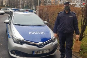 Dzielnicowy z Komisariatu II Policji w Bielsku-Białej idzie obok radiowozu podczas doręczania zakupów seniorce na kwarantannie