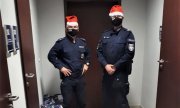 Zdjęcie przedstawia policjantów stojących na korytarzu komendy. Funkcjonariusze są w granatowych mundurach, a na głowach mają czerwone czapki. Obok nich stoją paczki z prezentami