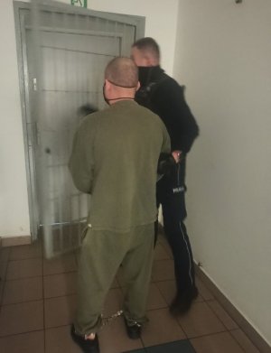 Na zdjęciu widoczny jest podejrzany, który prowadzony przez umundurowanego policjanta do wyjścia z pomieszczeń dla osób zatrzymanych.