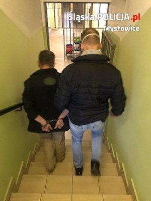 zatrzymany mężczyzna prowadzony przez nieumundurowanego policjanta korytarzem Pomieszczenia Dla Osób Zatrzymanych