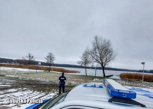 Dach policyjnego radiowozu, a w tle policjant sprawdzający czy na zamarzniętym jeziorze nie ma osób