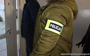 Na zdjęciu policjant w cywilnym ubraniu, z opaską na ręce z napisem policja prowadzi zatrzymanego mężczyznę korytarzem do aresztu