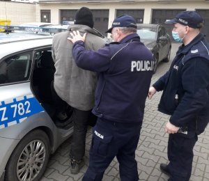 dwaj policjanci z zatrzymanym mężczyzną obok radiowozu policyjnego