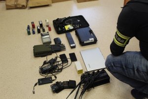 Policjant rozłożył na podłodze przedmiot zabezpieczone w trakcie przeszukania, takie jak laptopy, zagłuszarki, pieczątki, telefony
