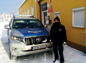 policjantka stoi przy radiowozie, który jest zaparkowany przed budynkiem posterunku Policji