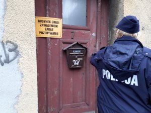 policjantka stoi przed budynkiem, na drzwiach wisi informacja: budynek grozi zawaleniem, zakaz przebywania