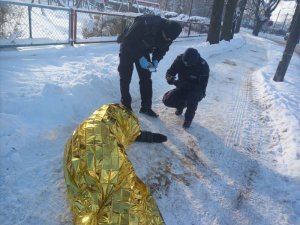 Zdjęcie przedstawia policjantów udzielających pomocy osobie potrzebującej. Osoba leży na chodniku, jest przykryta kocem termicznym. Na ziemi leży śnieg