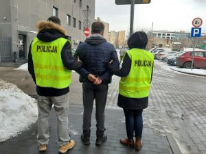 Zdjęcie jest zrobione na dziedzińcu Komendy Miejskiej Policji w Łodzi, widać na nim zatrzymanego ubranego w szare spodnie dresowe oraz ciemną kurtkę, na rękach ma założone kajdanki, po obu stronach trzymają go policjanci w zielonych kamizelkach policyjnych. Zdjęcie zrobione jest od tyłu