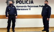 dwaj policjanci, którzy pomagali w ratowaniu dziecka stoją na tle ściany z napisem Policja Komenda Rejonowa Policji Warszawa III