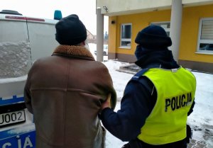 Umundurowany policjant prowadzi zatrzymanego. W tle oznakowany radiowóz oraz budynek KPP w Wieruszowie