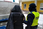 Umundurowany policjant prowadzi zatrzymaną. W tle oznakowany radiowóz oraz budynek KPP w Wieruszowie