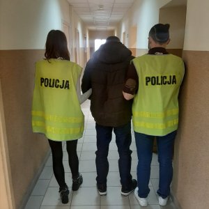 Policjant wraz z policjantką, ubrani w żółte kamizelki odblaskowe z napisem Policja, prowadzą przez korytarz w jednostce Policji zatrzymanego mężczyznę. Mężczyzna ubrany jest w brązową kurtkę