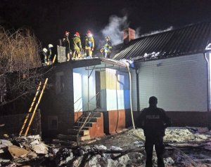 Stojący przed domem policjant, na dachu domu strażacy