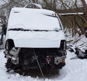 Na zdjęciu samochód typu bus koloru białego uszkodzony zasypany śniegiem. Pojazd nie posiada przednich elementów karoserii widać uszkodzony silnik