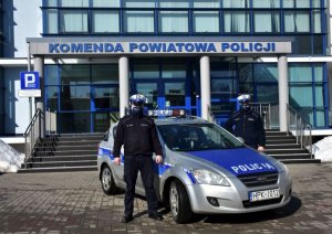 Policjanci wydziału ruchu drogowego stoją obok radiowozu. W tle budynek Komendy Powiatowej Policji w Jarosławiu